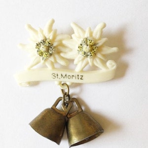 May include: White plastic brooch with two edelweiss flowers and a banner that reads "St. Moritz". The brooch has two small gold bells hanging from it.