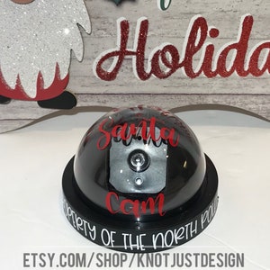 May include: A black dome-shaped Santa Cam doorbell with the text "Santa Cam" and "Property of the North Pole" in white lettering.