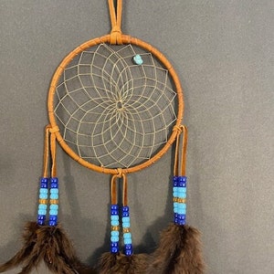 May include: A brown leather dreamcatcher with blue and gold beads and brown feathers. The dreamcatcher has a turquoise stone in the center.