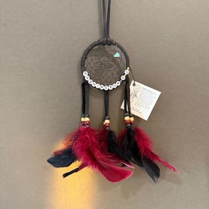 May include: Black dreamcatcher with red and black feathers. The dreamcatcher has the word "WASHINGTON D.C." spelled out in white beads. A small tag with text is attached to the dreamcatcher.