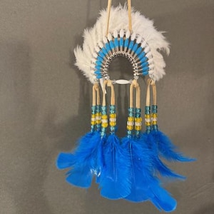 May include: A decorative Native American headdress featuring white feathers, a blue and white beaded band, and blue feather accents. The headdress is suspended by a leather cord.