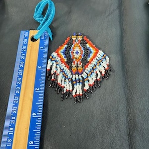May include: A beaded pendant with a geometric design in black, red, orange, white, and blue. The pendant is attached to a blue suede cord. A ruler is next to the pendant, showing measurements in inches.