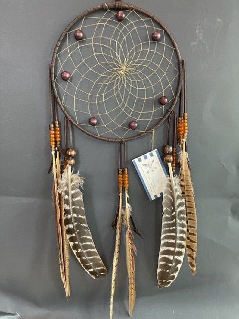 May include: A brown dreamcatcher with a beige net, brown leather accents, and purple beads. It has a white tag with the text "Dreamcatchers by" and a logo. It has brown and white feathers hanging from the bottom.