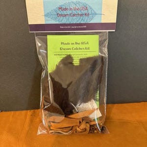 May include: A clear plastic bag containing a dream catcher kit. The kit includes brown feathers, tan leather strips, and beads. The packaging features text that reads "Made in the USA Dream Catcher Kit."