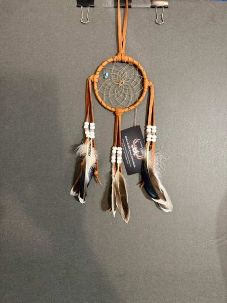 May include: A tan leather dreamcatcher featuring a circular hoop with a woven web, a single turquoise bead, and three hanging leather strips. Each strip is adorned with white beads and natural brown and white feathers. A black tag is attached.