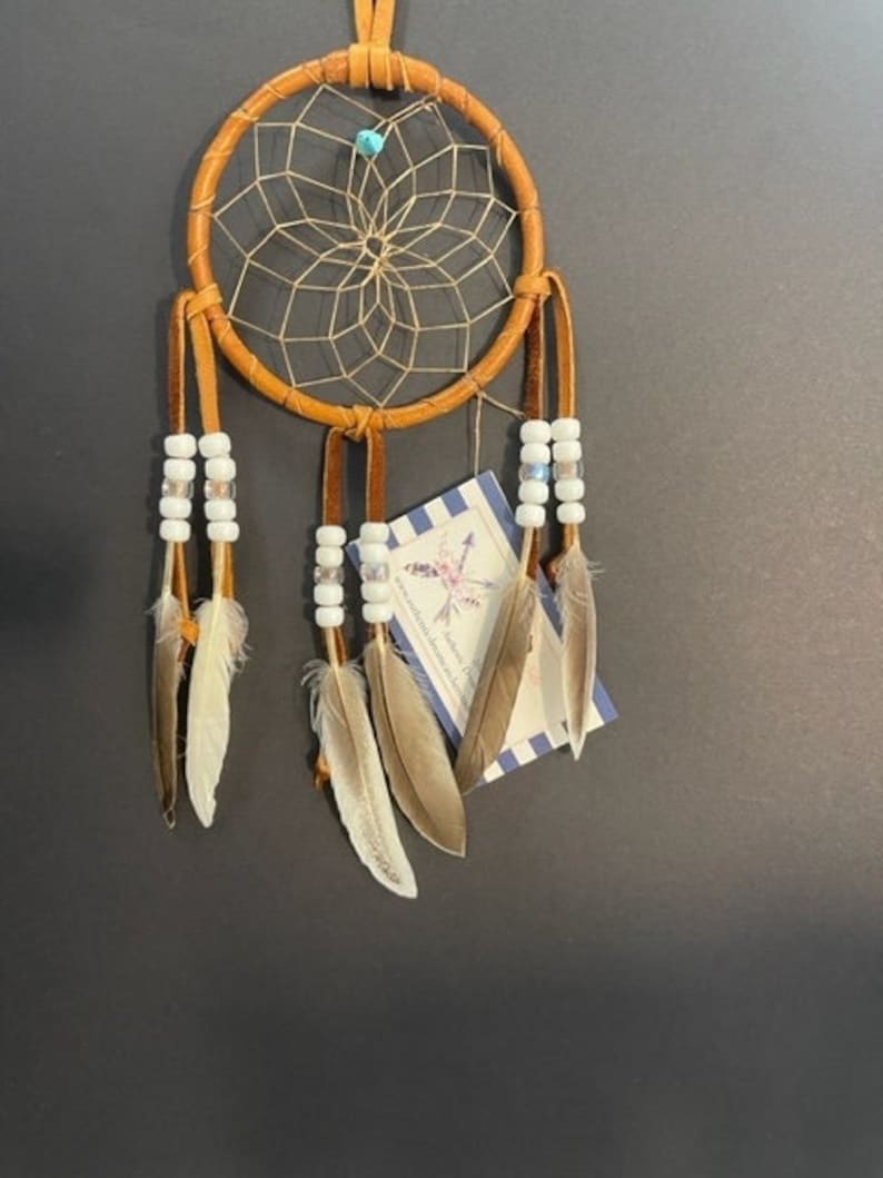 May include: A circular dream catcher wrapped in tan leather with a woven web center featuring a small turquoise bead. Three leather strips hang from the bottom, adorned with white beads and natural brown and white feathers. A tag is attached.