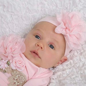 May include: A baby wearing a pink outfit and a matching headband with a large flower. The baby is lying on a white, textured surface. The outfit has floral and lace embellishments.