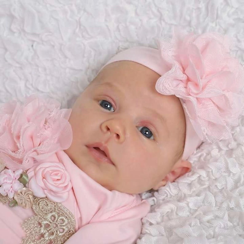 May include: A baby wearing a pink outfit and a matching headband with a large flower. The baby is lying on a white, textured surface. The outfit has floral and lace embellishments.