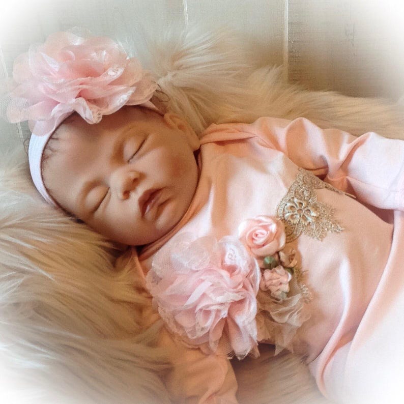 May include: A sleeping baby wearing a light pink onesie with floral embellishments and a matching headband with a large pink flower. The baby is lying on a fluffy, white surface.