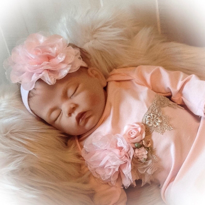 May include: A sleeping baby doll wearing a light pink outfit with a matching floral headband. The outfit features a decorative floral applique and is lying on a fluffy, white surface.