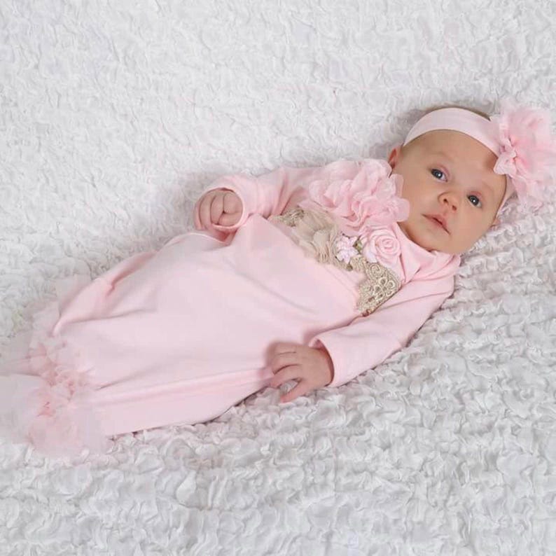 May include: A baby in a light pink gown and matching headband with a large flower. The gown has long sleeves and a ruffled hem. A large pink flower and lace embellish the chest area. The baby is lying on a white, textured surface.