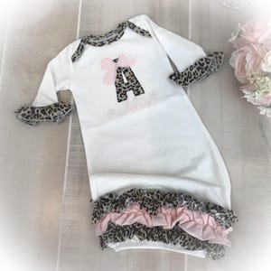 May include: White baby gown with leopard print accents. Features a pink bow and the letter "A" in a leopard print design. The name "Caroline" is embroidered in pink script. Ruffled trim at the sleeves and hem.
