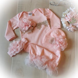 May include: A baby clothing set in soft pink, including a long-sleeved onesie, a hat, a headband, and a footed sleep sack. The onesie features a floral applique and lace detail. The sleep sack has a ruffled trim. The set is designed for newborns.