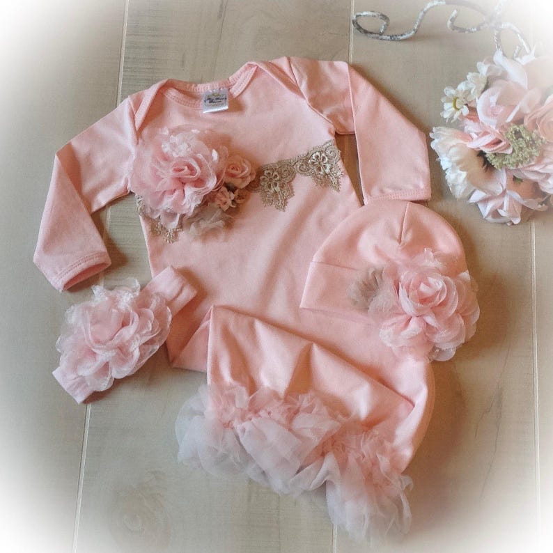 May include: A baby clothing set in soft pink, including a long-sleeved onesie, a hat, a headband, and a footed sleep sack. The onesie features a floral applique and lace detail. The sleep sack has a ruffled trim. The set is designed for newborns.