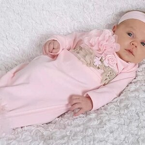 May include: A baby wearing a light pink gown and matching headband with a large flower. The gown has long sleeves and a ruffled hem. The outfit is decorated with lace and additional flowers.