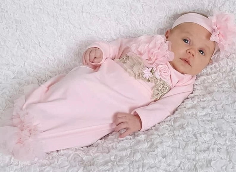 May include: A baby wearing a light pink gown and matching headband with a large flower. The gown has long sleeves and a ruffled hem. The outfit is decorated with lace and additional flowers.