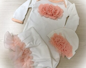 May include: A white baby gown with long sleeves and a peach-colored flower embellishment on the chest. The gown is paired with a matching peach-colored flower hat.