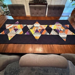 May include: A Halloween-themed table runner with star-shaped patchwork designs. The runner features a black background with orange, purple, yellow, and white patterned star shapes. The runner is placed on a wooden table.