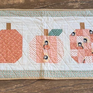 May include: A rectangular quilted table runner featuring five pumpkin shapes in various shades of orange and peach. The pumpkins are set against a cream background with a green and white checkered border. Each pumpkin has a brown stem and green leaf.