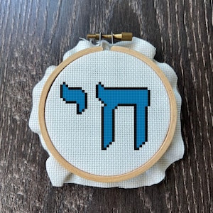 May include: A circular wooden embroidery hoop containing white Aida fabric with a cross-stitched design of the Hebrew word Chai in bright blue thread. The design features the two Hebrew letters Chet and Yod centred within the frame.