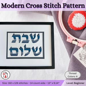 May include: A framed cross-stitch pattern with the words "Shabbat Shalom" in blue and white. The black frame surrounds the artwork, which is on a white background. A pink embroidery hoop and a red and white stitched circle are visible.