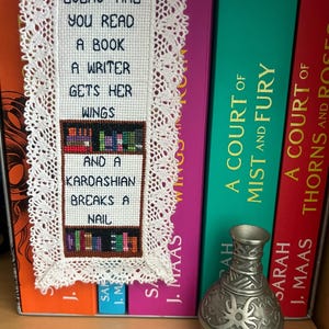 Könnte beinhalten: Ein weißer Kreuzstich-Lesezeichen mit dem Text "Every time you read a book a writer gets her wings and a Kardashian breaks a nail". Das Lesezeichen ist mit weißer Spitze umrahmt und wird in einem Bücherregal mit mehreren Büchern ausgestellt. Die Bücher sind alle von Sarah J. Maas und tragen die Titel "A Court of Mist and Fury" und "A Court of Thorns and Roses".