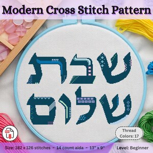 May include: A blue and white cross stitch pattern with the Hebrew words "Shabbat Shalom" in a modern style. The pattern is 13 inches by 9 inches and uses 17 colors of thread.