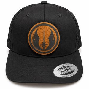 Puede incluir: Una gorra snapback negra con un parche circular marrón de textura desgastada y un símbolo Jedi negro. La gorra tiene una parte trasera de malla y una pegatina plateada en la visera que dice THE AUTHENTIC YUPOONG SNAPBACK YUPOONG SINCE 1974.