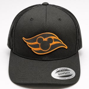 Puede incluir: Una gorra snapback negra con parte trasera de malla y un parche frontal con acabado de cuero marrón que muestra un icono estilizado de Mickey Mouse. Una pegatina plateada y negra en la visera dice THE AUTHENTIC YUPOONG SNAPBACK YUPOONG SINCE 1974.