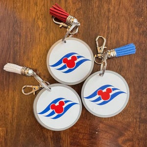 May include: Three round keychains with a silver metal ring and a colorful tassel. Each keychain features a white circle with a blue and red Mickey Mouse logo. The logo is a stylized Mickey Mouse head with three blue waves.