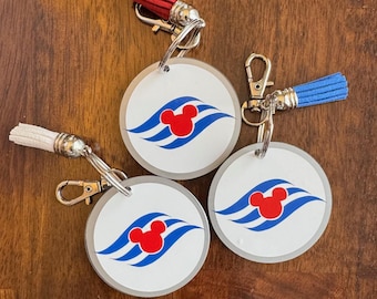 May include: Three round keychains with a silver metal ring and a colorful tassel. Each keychain features a white circle with a blue and red Mickey Mouse logo. The logo is a stylized Mickey Mouse head with three blue waves.