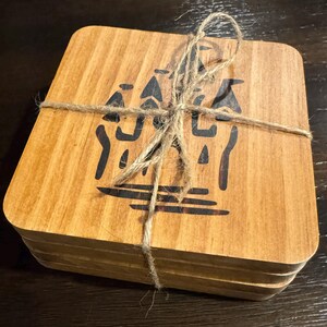 May include: A stack of square wooden coasters with a dark brown graphic design and tied with twine. The coasters have a light brown wood grain pattern and are stacked neatly. The graphic design appears to be an abstract representation of a building.