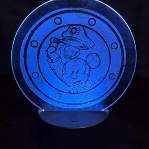 May include: A blue-lit, circular night light featuring a sketch of Mickey Mouse in a hat. The design is etched into a clear disc, surrounded by a ring of circles. The base is black with a power symbol.