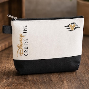 May include: A rectangular canvas pouch featuring a two-tone design with a white upper section and a black lower section. The text Disney Cruise Line is printed in gold glitter on the left, with a matching gold glitter Mickey Mouse icon logo.