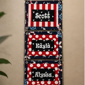 May include: A vertical hanging sign with five rectangular fabric panels connected by metal rings. The top panel is black with the text "Cruisin' with mickey" and anchor icons. The four lower panels feature red and white stripes or polka dots with black center labels displaying the names Scott, Kayla, Alyssa, and Zdek, accented with small white Mickey Mouse head silhouettes.