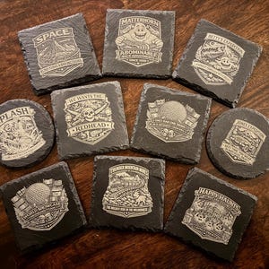 May include: A collection of black slate coasters with white engraved designs and text. The coasters feature various themes, including "SPACE", "SPLASH", and "HAPPY HAUNTS". The coasters are in various shapes, including square and round.