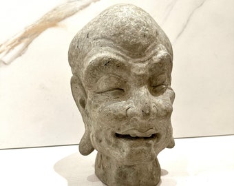 May include: A gray stone sculpture of a man's head with a closed-eye expression and a slight smile. The sculpture is detailed with wrinkles and a textured surface.