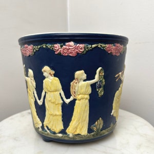 May include: A dark blue ceramic planter with a white interior and a decorative relief. The relief depicts figures in yellow gowns holding hands, with pink roses and green foliage around the rim. The planter has three small feet.