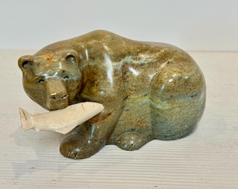 May include: A ceramic bear figurine in shades of brown, tan, and green, holding a cream-colored fish in its mouth. The bear is in a seated position, with detailed paws and a textured surface. A decorative item.