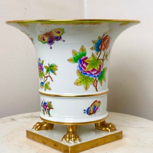 Könnte beinhalten: Eine weiße Porzellanvase mit goldenen Verzierungen und floralen und Schmetterlingsmotiven. Die Vase hat einen quadratischen Sockel mit vier goldenen Löwenpfoten.