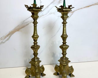 May include: A pair of antique brass candlesticks with ornate details and green accents on the top. The candlesticks have a tall, slender design with a square base and three legs.