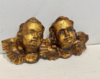 May include: Two gold cherub angel figurines with wings, each with a detailed face and hair. The cherubs are facing each other and are attached at the wings.