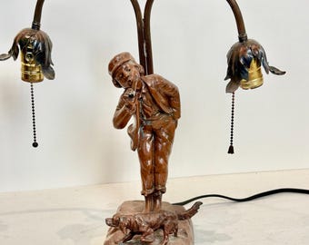 May include: A vintage bronze-toned table lamp featuring a sculpted figure of a boy with a dog. The lamp has two curved arms with floral-shaped shades and pull chains. The base is a textured, rectangular shape.