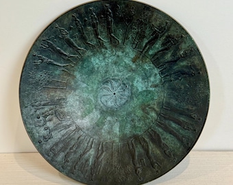 May include: A round, antique bronze plate with a detailed relief design. The plate features a central spiral pattern and a surrounding band of figures and trees. The bronze has a green patina, giving it an aged appearance.