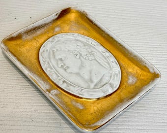 May include: A small, rectangular ceramic dish with a gold-colored glaze. The dish features a white, raised relief of a woman's profile with a floral wreath.