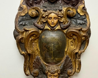 May include: Ornate, antique wooden wall plaque with a central painted scene. The plaque features a gold-colored frame with dark brown and black accents, and includes sculpted faces and angel wings.