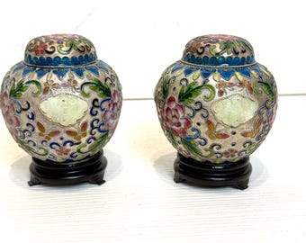 May include: Two small, round, decorative jars with lids. The jars are made of white porcelain with intricate floral designs in shades of pink, blue, green, and gold. The jars are sitting on black wooden bases.