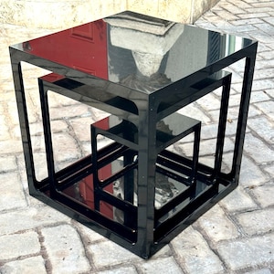 May include: A set of three nesting tables in a modern design. The tables are made of black acrylic with a glossy finish. The largest table is approximately 24 inches square, the middle table is 18 inches square, and the smallest is 12 inches square.