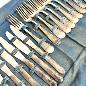 Puede incluir: Un juego de cubiertos de plata en un estuche de terciopelo azul. Los cubiertos están dispuestos en filas e incluyen tenedores, cuchillos y cucharas. El estuche tiene un forro de rayas azules y blancas. Los cubiertos están grabados con las palabras "First National Bank of New York" y "The First National Bank of New York".