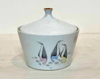 May include: A white ceramic lidded container with a gold-trimmed finial. The container features three stylized penguins with black, white, and colorful accents. The lid has a textured, ridged design, and the container has a light blue background.